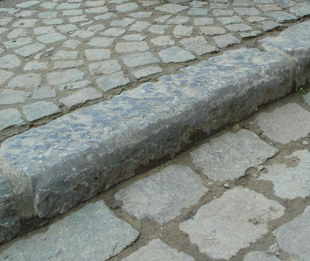 Prior to the abundant use of concrete as the standard material, curbs were made of local quarried or other mined stone materials.  Granite, such as seen above, made an excellent (although expensive) material for curbing, as its hardened nature reduced its overall susceptibility to erosion and damage from street activities (e.g. cart and horse traffic, non-inflatable wheels, pedestrians).  Its weight and density also was an advantage over softer stones as it made a rigid edge that was easy to clean and hard to move or steal.   Photo rackcdn.com